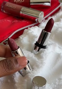 To KIKO-leppestifter i sølvfargede tuber, en burgunder og en med mønster, delvis begravet i hvit snø ved siden av en rød pose.