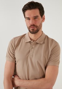 Bež polo majica s teksturiranim, saćastim uzorkom. Placket s gumbima, kratki rukavi i klasičan ovratnik. Glatka tkanina, opušteni dizajn.