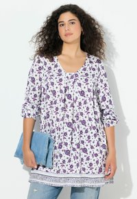 Tunika floreale con scollo a V profondo, in bianco e viola, con maniche lunghe, dettagli arricciati e orlo decorato. Tiene in mano un capo in denim blu piegato.