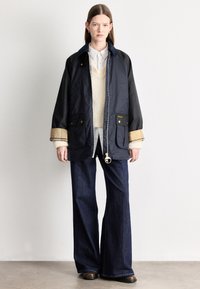 Tengerészkék dzseki hegyes gallérral, két első zsebbel és cipzáros rögzítéssel, világos pulóver fölött viselve, bővülő sötét farmerrel párosítva.