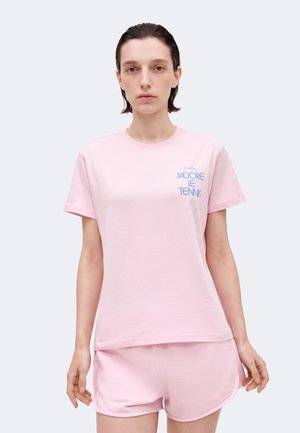 Γυναίκα με κοντά σκούρα μαλλιά, που φοράει ανοιχτό ροζ μπλουζάκι με μπλε κείμενο "J'adore le Tennis" και ροζ σορτς που ταιριάζουν, σε απλό φόντο.