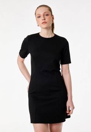 Młoda kobieta w dopasowanej czarnej sukience z krótkim rękawem, stojąca z jedną ręką za plecami i neutralnym wyrazem twarzy, na jednolitym białym tle.