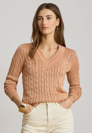 Femme portant un pull à col en V en maille torsadée couleur pêche et un pantalon crème, les mains dans les poches et un bracelet manchette en or au poignet gauche.