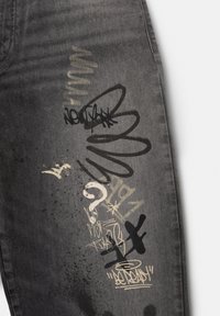Jambe de jean en denim noir avec des inscriptions superposées blanches et noires de style graffiti, des gribouillis, un point d'interrogation et des éclaboussures de peinture sur le tissu.