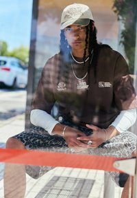 Brown oversized t-shirt with white long sleeves, leopard print shorts, beige cap, layered necklaces, and silver rings. Sitting pose.