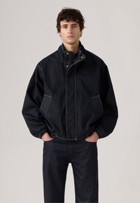 Donker denim jack met een hoge kraag, een ritssluiting aan de voorkant en twee zijzakken. Contraststeek en elastische manchetten voegen detail toe.
