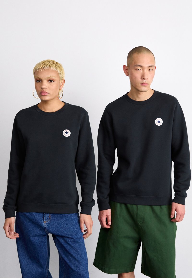 Los sudaderas de cuello redondo negras cuentan con un parche de logo en el pecho. Un modelo lleva vaqueros azules sueltos, el otro lleva pantalones cortos verdes. Diseño minimalista, textura suave.