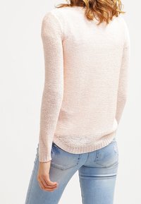 Pull en tricot rose clair à manches longues, motif texturé et ourlet légèrement arrondi. Porté avec un jean bleu clair déchiré.
