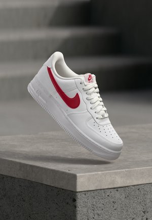 Baskets blanches Nike Air Force 1 avec logo swoosh rouge flottant au-dessus d'un rebord en béton et des escaliers en arrière-plan.
