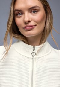 Junge Frau mit hellem Haar, die ein weißes, strukturiertes Oberteil mit Reißverschluss und rundem Zipperanhänger trägt, ruhiger Gesichtsausdruck, neutraler grauer Hintergrund.