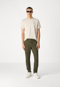 T-shirt beige clair, col rond, avec un petit logo ; pantalon ajusté vert olive ; baskets blanches avec des accents noirs ; lunettes de soleil.