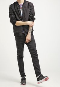 Black bomber jacket with ribbed cuffs, black distressed jeans, and black athletic shoes with red accents. Colorful graphic t-shirt underneath.