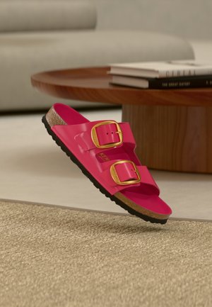 Roosad plastist sandaalid kahe kuldse pandlaga, tekstuuriga tallaga ja musta kummist tallaga, hõljuvad tekstuuriga beeži pinna kohal.