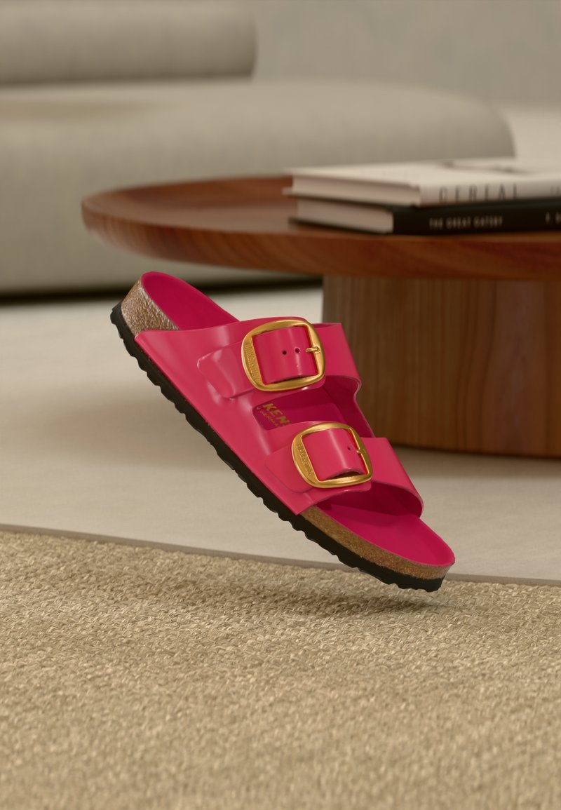 Ροζ πλαστικά σανδάλια με δύο χρυσά αγκράφες, υφασμένο πέλμα και μαύρη λαστιχένια σόλα, να αιωρούνται πάνω από μία υφασμένη μπεζ επιφάνεια.