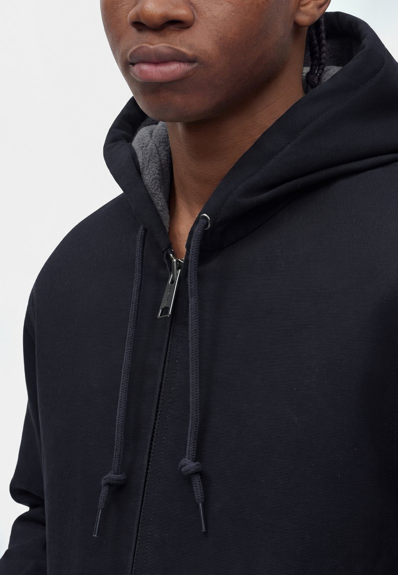 Primer plano de una persona que lleva una sudadera con capucha negra con cremallera y cordones, mostrando la parte inferior de la cara, el cuello y la parte superior del pecho.