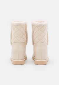 Stivali invernali beige imbottiti con morbido rivestimento in shearling piegato sulla parte superiore, visti da dietro su uno sfondo bianco.