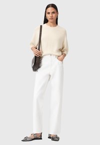 Maglione a maglia crema, pantaloni bianchi a gamba dritta, scarpe grigie flat con punta affusolata. Modella con borsa a tracolla marrone, accessori minimi.