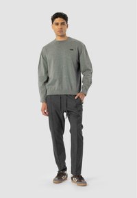 Hombre de pie que lleva un suéter gris de cuello redondo, pantalones oscuros de tablones y zapatillas Adidas marrones, frente a un fondo claro y liso.