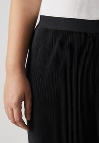 Black pleated trousers with a wide elastic waistband, featuring vertical lines and a soft texture. Hand visible at the edge of the frame.