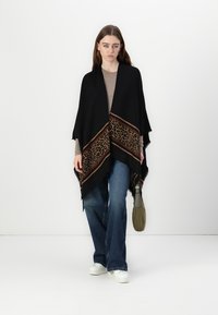 Poncho nero con fantasia leopardata e strisce diagonali marroni, con bordi frangiati. Abbinato a un maglione leggero e jeans.