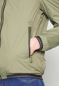Chaqueta bomber verde oliva con cierre de cremallera, puños elásticos y un bolsillo lateral. Presenta una textura suave y acentos negros contrastantes.