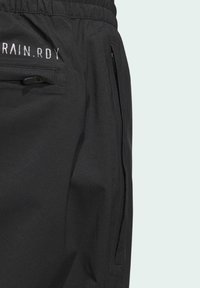Un pantalon noir imperméable avec un tissu texturé, une ceinture élastique et une poche zippée sur le côté avec le texte "RAIN.RDY" sur la hanche.