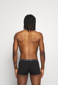 Schwarze Baumwoll-Boxershorts mit einem weichen Bund, der mit dem Markenlogo versehen ist. Das Modell zeigt einen glatten Rücken mit sichtbarer Hautstruktur und Tätowierungen.