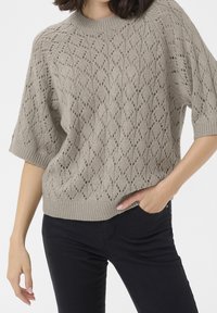 Personne portant un pull en tricot beige à manches courtes avec des motifs en losange et un pantalon noir, une main dans la poche, sur un fond blanc.