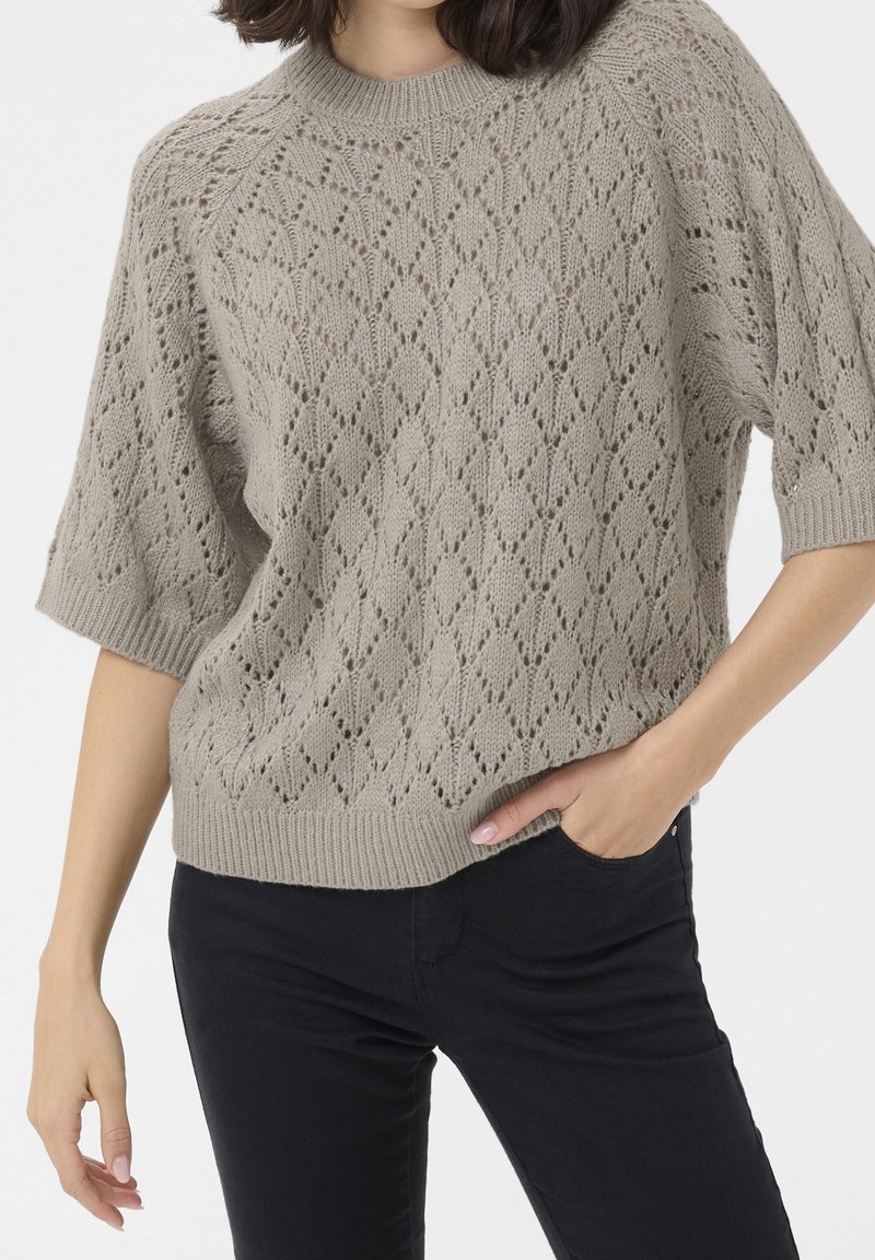 Personne portant un pull en tricot beige à manches courtes avec des motifs en losange et un pantalon noir, une main dans la poche, sur un fond blanc.