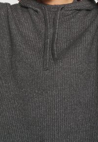 Nahaufnahme einer Person, die einen dunkelgrauen gestrickten Kapuzenpullover trägt, mit sichtbarem strukturiertem Stoff und hängenden Zugkordeln.