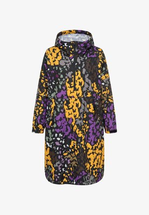 Femme portant un imperméable oversize coloré à motifs avec des éclaboussures violettes, jaunes et noires, associé à un jean bleu et des bottes grises montant jusqu’aux genoux.
