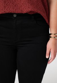 Hoge taille zwarte jeans met een gestructureerde tailleband, voorzakken en subtiele stiksels. Gecombineerd met een bordeauxrood top en gouden armbanden.