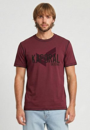 Homme blond avec une barbe portant un t-shirt Kaporal Original bordeaux et un jean bleu clair, debout devant un fond uni.