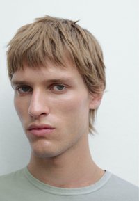 Close-up of a person with short, light brown hair, wearing a soft, light green t-shirt. Clear skin and neutral expression, against a plain background.