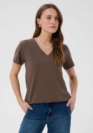 Frau mit langen braunen Haaren, die ein braunes V-Ausschnitt-T-Shirt, blaue Jeans, eine silberne Halskette und Ohrringe trägt, steht mit den Händen in den Taschen vor weißem Hintergrund.