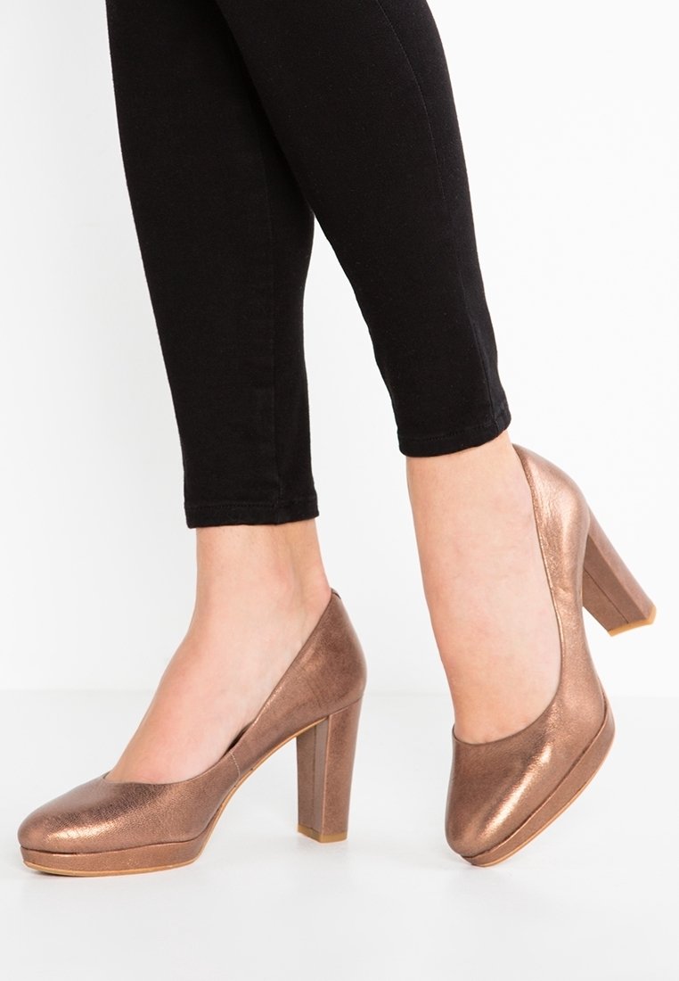 Femme portant des escarpins métalliques bronze à talons hauts carrés avec un pantalon noir ajusté, sur fond blanc.