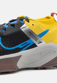 Chaussure de trail running noire et jaune avec des accents bleus, une tige en mesh texturé et une semelle rembourrée arborant un design Nike swoosh bien visible.