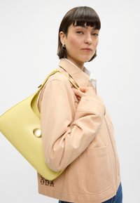 Φωτεινό ροζ ζακέτα με φερμουάρ και μπροστινές τσέπες, διακοσμημένη με καφέ κεντήματα, συνδυασμένη με μια κίτρινη τσάντα χεριού με στρογγυλό κούμπωμα και λεία υφή.