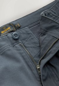 Tmavě šedé kalhoty vyrobené z odolné tkaniny, které mají zipový klíč, knoflíkové zapínání a štítek s nápisem "NEWM Durable Goods."