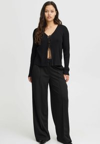 Svart ribbad cardigan med långa ärmar, V-hals och framslipsar. Kombinerad med svarta vida byxor, vilket skapar en strömlinjeformad silhuett.