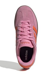 Rosa mockaskin Adidas-sneakers med orangea ränder och snören, svart innersula med vitt Adidas-logotyp, sedd uppifrån mot vit bakgrund.
