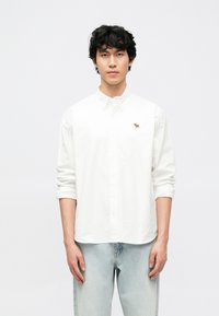 Λευκό πουκάμισο με κουμπιά από βαμβάκι, με μικρή κεντητή καφέ εικόνα ελάφι στ' αριστερό στήθος. Το μοντέλο φοράει ανοιχτό μπλε τζιν παντελόνι.