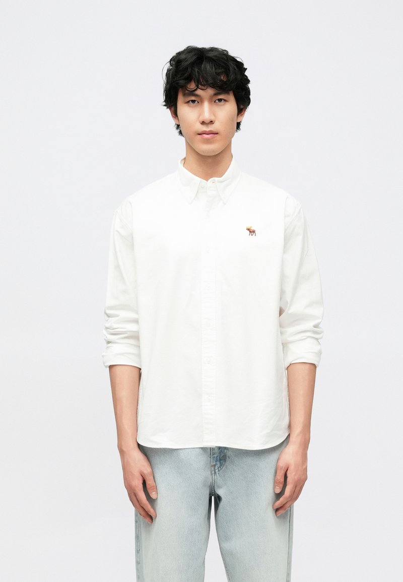 Λευκό πουκάμισο με κουμπιά από βαμβάκι, με μικρή κεντητή καφέ εικόνα ελάφι στ' αριστερό στήθος. Το μοντέλο φοράει ανοιχτό μπλε τζιν παντελόνι.