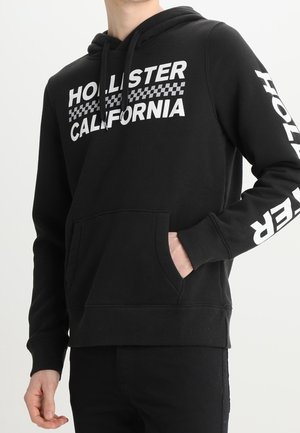 Osoba ubrana w czarną bluzę z kapturem z białym napisem "Hollister California" oraz wzorem w kratkę na piersi i rękawie, z ręką w przedniej kieszeni.