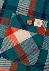 Camicia di cotone a scacchi con un motivo turchese, rosso e blu scuro, una tasca sul petto con un bottone di legno e un'etichetta in tessuto con un accento di design.