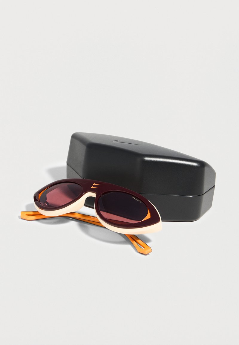 Gafas de sol ovaladas con lentes negras y montura en tonos marrón, crema y naranja, descansando frente a un estuche rígido negro cerrado sobre una superficie blanca.