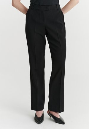 Personne portant un pantalon noir droit sur mesure et des chaussures noires à talons bas pointus sur fond blanc.