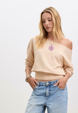 Maglione beige a spalle scoperte con orlo a coste, indossato con jeans a vita alta blu chiaro. La modella indossa una collana floreale viola e braccialetti sovrapposti.