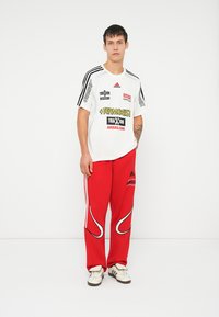 Bílý krátký rukáv tričko s černými a žlutými logy; červené sportovní kalhoty s bílými akcenty; model v bílých teniskách.