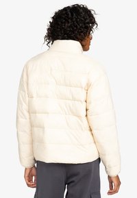 Puffjacka i krämfärgad med horisontell sömnad, hög krage och elastiska ärmar. Texturerat, lätt material som är lämpligt för kallt väder.